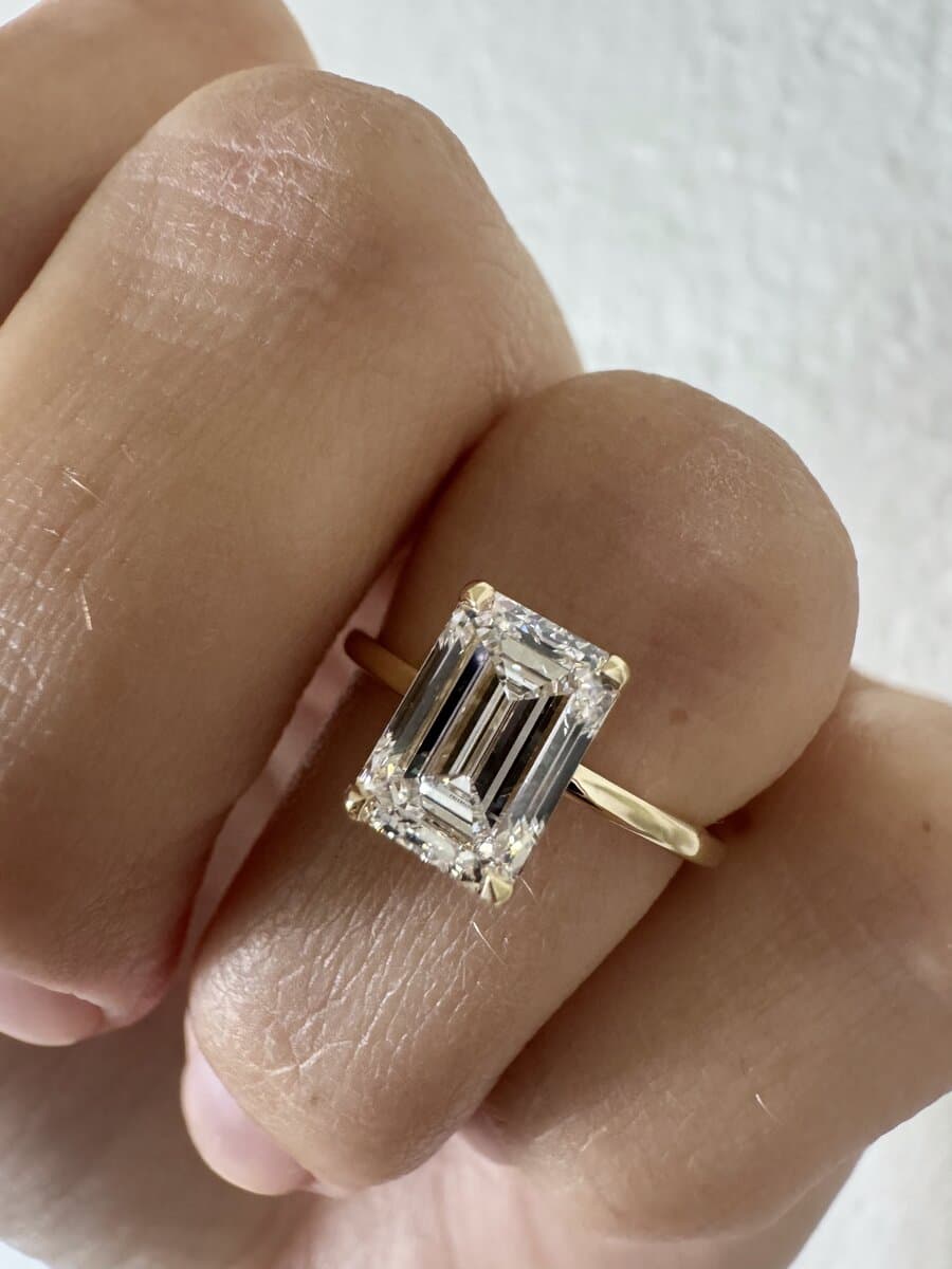 An emerald-cut diamond solitaire in yellow gold, worn on a hand in sunlight