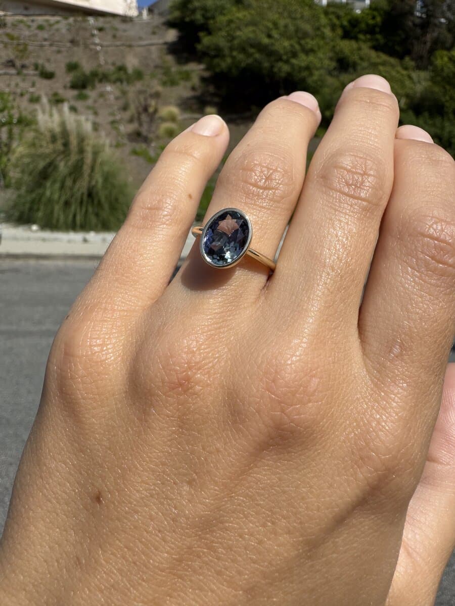 A hand wearing a blue-gray oval stone in a yellow gold bezel ring, outdoors