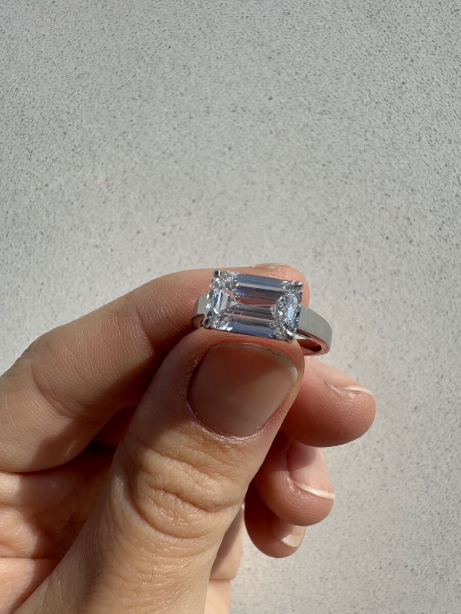 An east-west emerald-cut diamond ring in white metal, held between two fingers