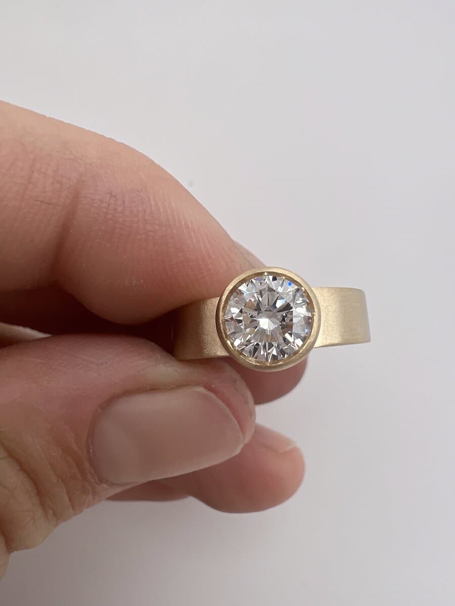 A round diamond in a wide yellow gold bezel band, held between two fingers