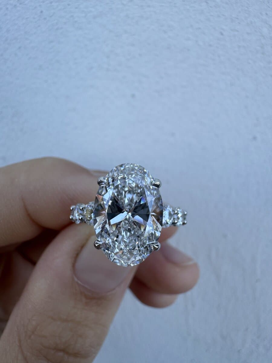 An oval diamond with pear-shaped side stones in a white-metal ring, held between two fingers