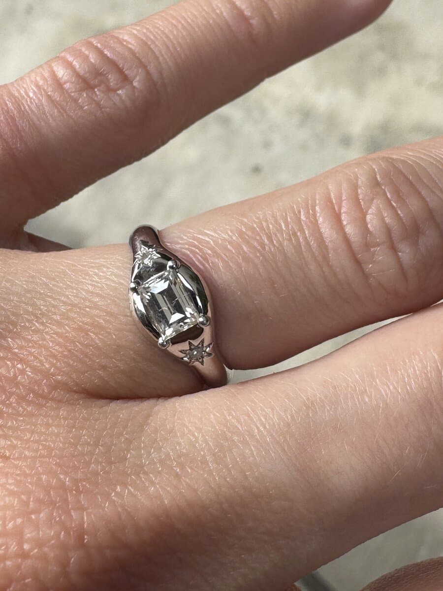 A hand wearing an emerald-cut diamond in a white-metal setting with a small side stone