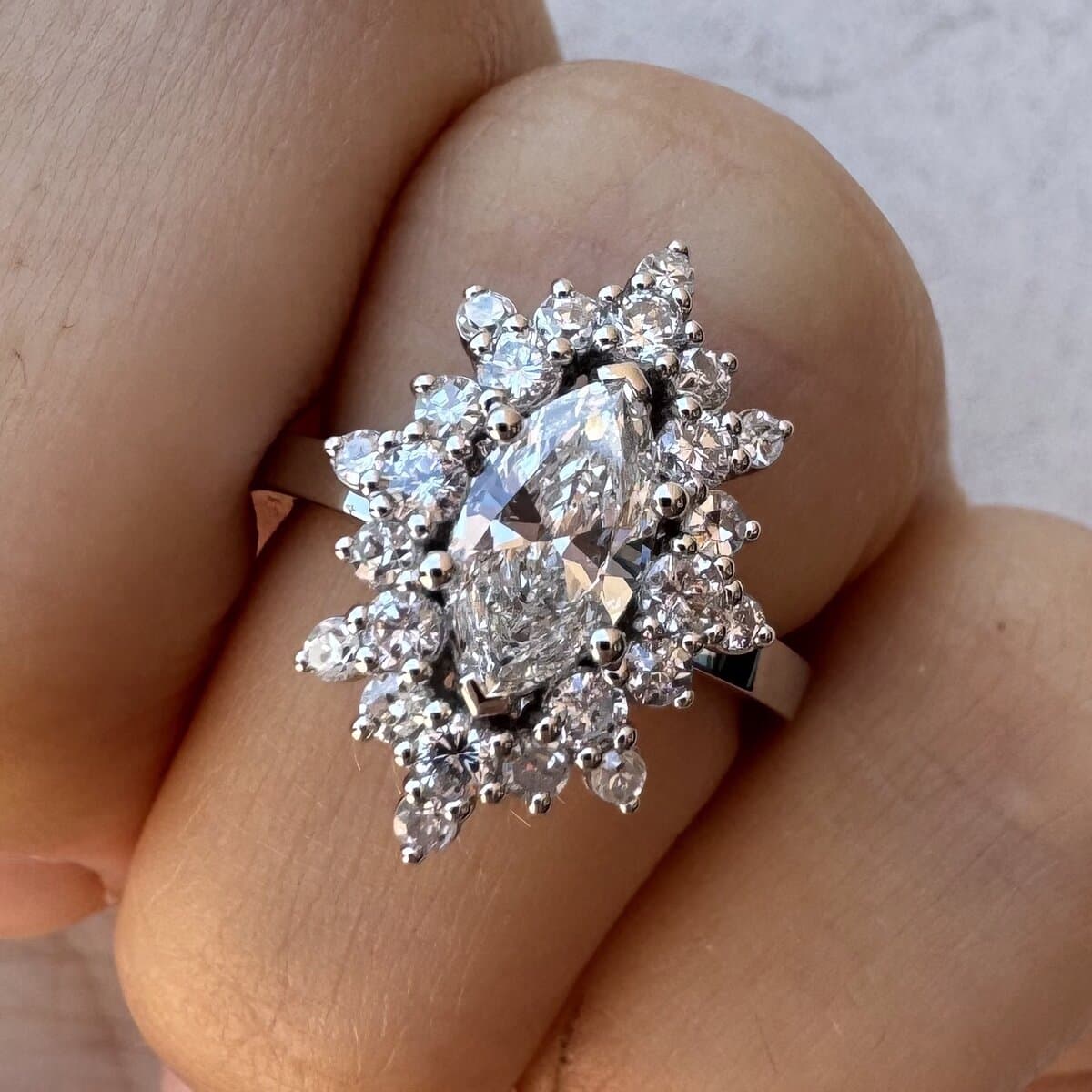 A marquise diamond cluster ring with a scalloped halo in white metal, worn on a hand