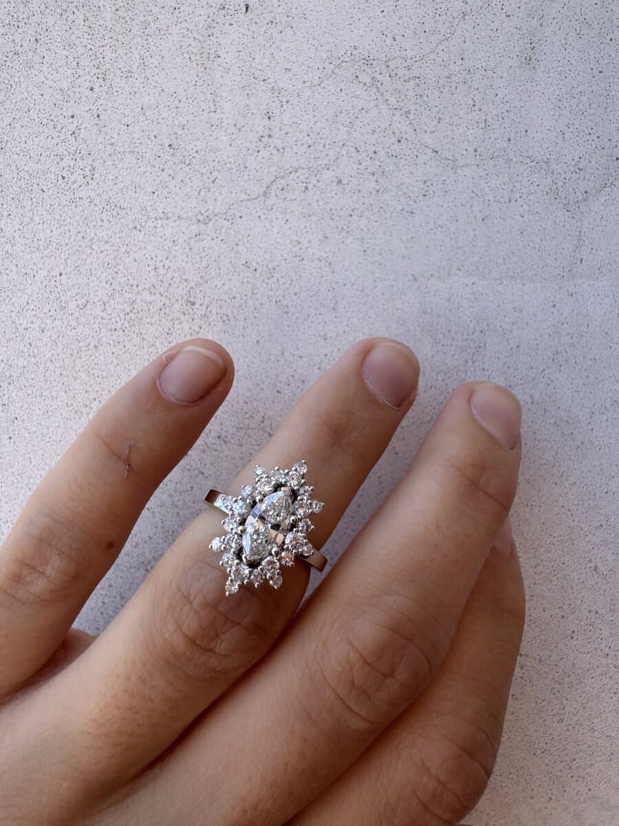 A hand wearing a marquise diamond cluster ring in white metal, against a white wall