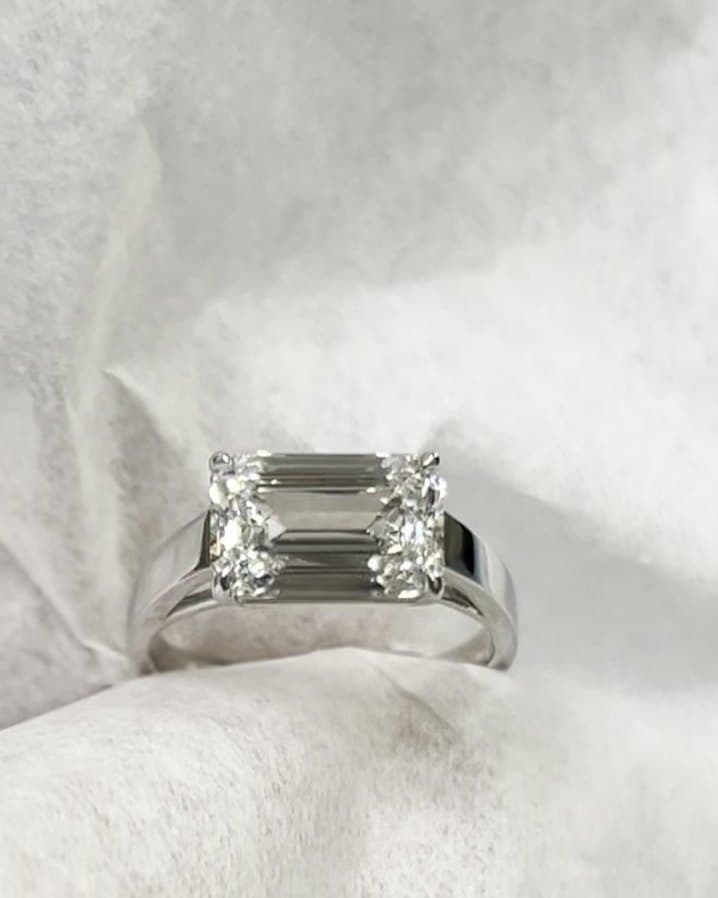 An east-west emerald-cut diamond ring in white metal, resting on white tissue