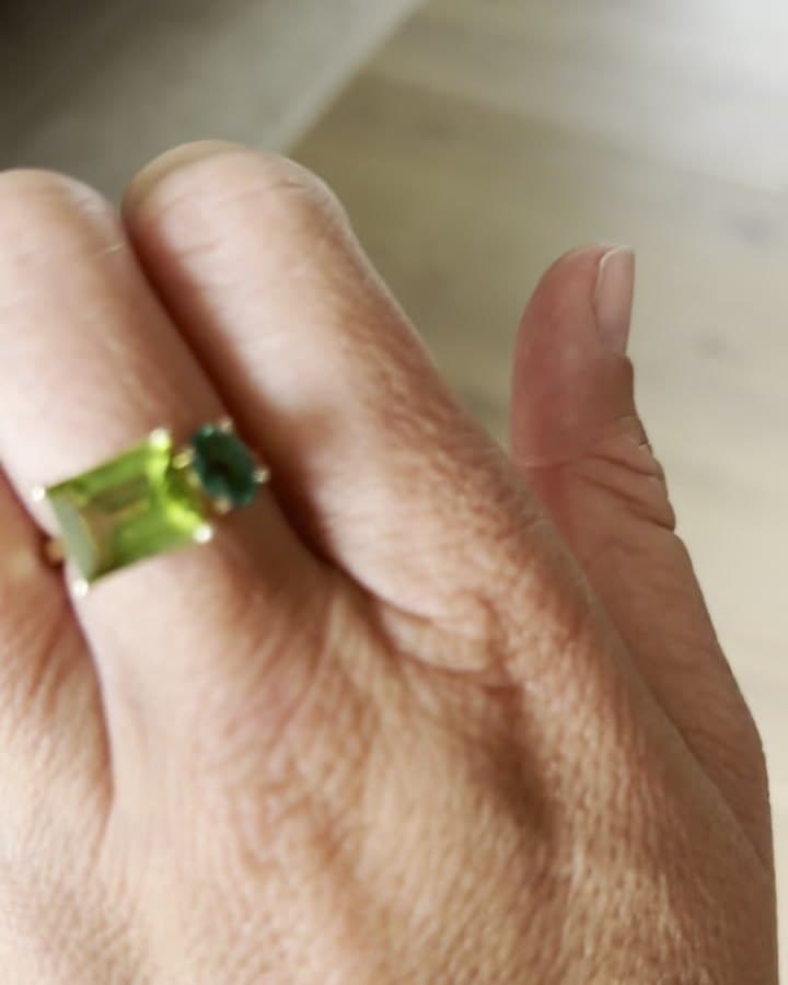 A yellow gold toi-et-moi ring with a green emerald-cut stone beside a smaller green round stone, worn on a hand