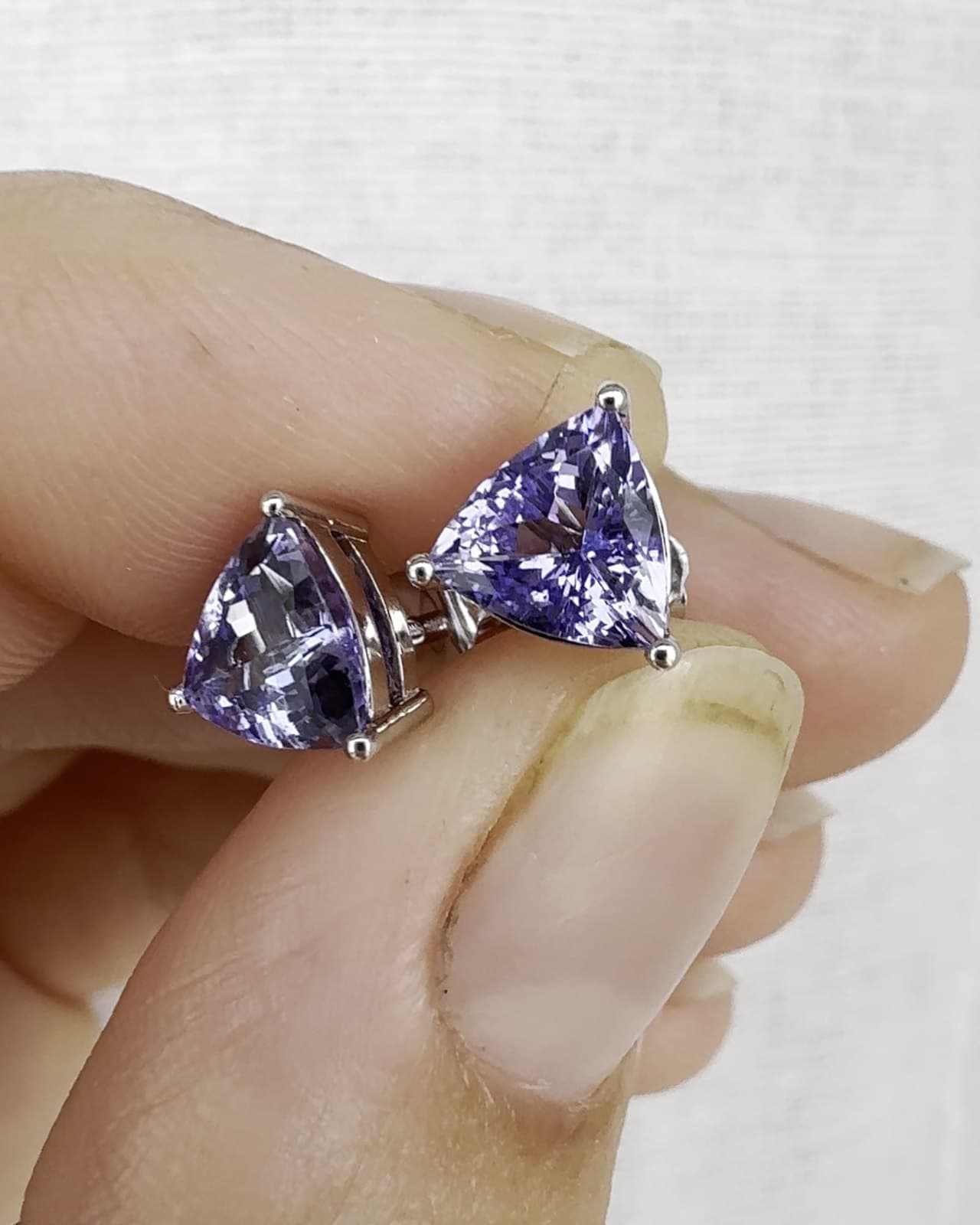 A pair of violet trillion-cut gemstones in white metal settings, held between two fingers